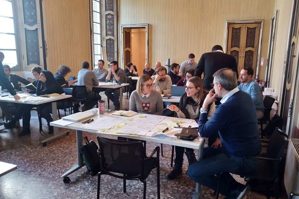 Corsisti divisi in gruppi lavorano ai tavoli in una sala con soffitti alti e porte decorate
